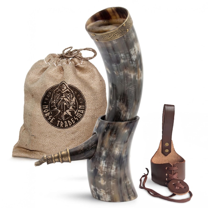 May include: A drinking horn with a polished, mottled grey and black finish, featuring a brass rim with an etched knotwork pattern. It includes a brown leather belt holster with lace-up details and a tan burlap drawstring bag labeled Norse Tradesman.