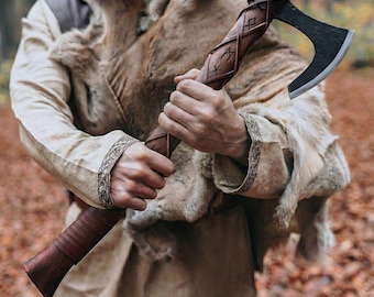 Può includere: Un'accetta con testa in metallo scuro e manico in legno avvolto da strisce di cuoio marrone. Il manico presenta simboli runici intagliati ed è tenuto da una persona che indossa una tunica beige con polsini ricamati e pelliccia marrone.