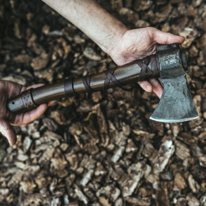 May include: A hand-forged axe featuring a dark, textured metal head and a smooth, cylindrical wooden handle. The handle is wrapped with thin, dark brown leather strips in a crisscross pattern, with additional leather bands securing the grip.