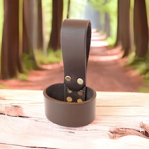 May include: A brown leather belt loop with a metal buckle. The loop is designed to hold a belt or strap.