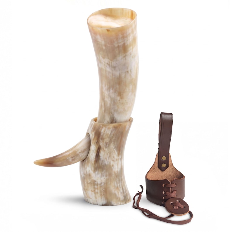 May include: A polished drinking horn with a natural cream, tan, and brown marbled pattern, featuring a smaller horn-shaped handle attached to the side. Beside it sits a dark brown leather belt holster with a lace-up design and a button closure.