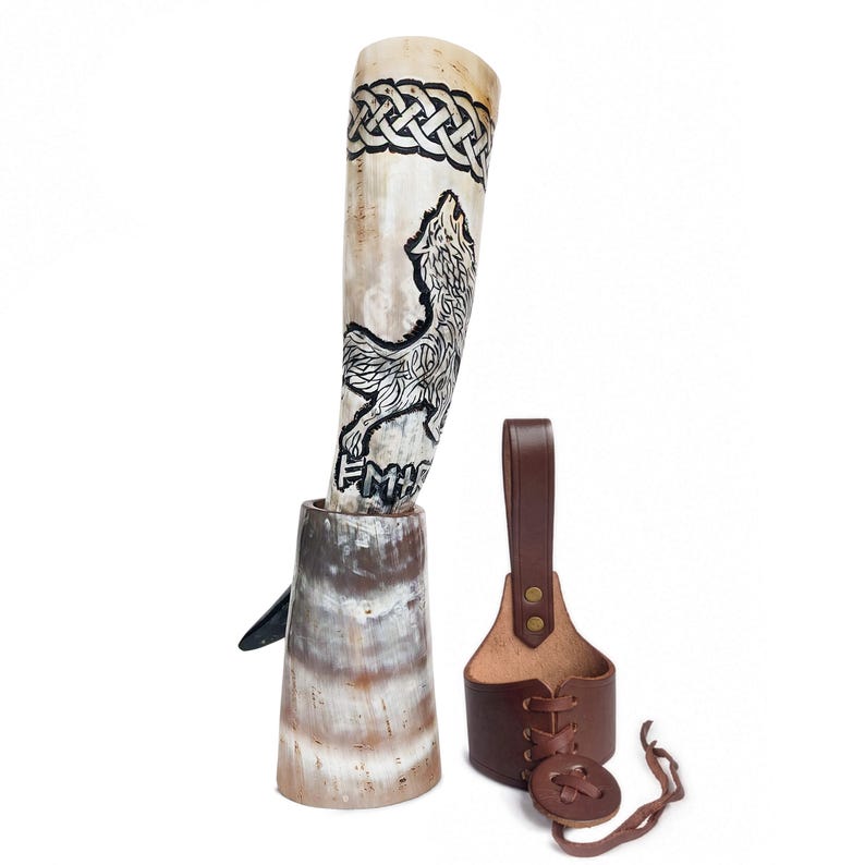 May include: A drinking horn with a cream-colored surface featuring a black etched wolf design, a Celtic knot pattern at the top, and runic characters. It is paired with a brown leather holder featuring a lace-up front and a vertical strap attachment.