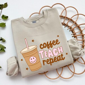 Op de afbeelding: Beige sweatshirt met een bruine en roze afbeelding van een koffiebeker met een smiley en de tekst "coffee teach repeat".
