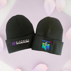 May include: Two black knit beanies with embroidered logos. One beanie has a purple, blue, and white Nintendo GameCube logo. The other beanie has a multi-colored Nintendo 64 logo.
