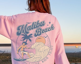 May include: Light pink sweatshirt with a Malibu Beach graphic on the back. The design features a surfer riding a wave, a palm tree, and the words "RIDE TIGHT, HANG LOOSE". The sweatshirt is a soft, comfortable material.