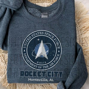 Könnte beinhalten: Ein dunkelblaues Sweatshirt mit dem Logo der United States Space Force. Das kreisförmige Logo zeigt einen Stern, einen Globus und den Text "ROCKET CITY Huntsville, AL". Das Sweatshirt liegt auf einer gewebten Matte.
