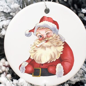 May include: A white ceramic Christmas bauble depicting a cartoon Santa Claus wearing a red hat and suit, winking and holding a cigarette in his mouth.