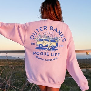 May include: A light pink sweatshirt with a graphic print of a van with a surfboard on top. The graphic also includes the text "Outer Banks", "Sand", "Surf", "Pogue Life", and "North Carolina".
