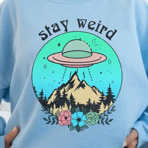 May include: Light blue sweatshirt with a graphic of a UFO flying over a mountain range with a forest and flowers. The text "stay weird" is above the graphic.