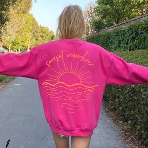 May include: A pink sweatshirt with a graphic of a sunset over the ocean and the text "sunset wanderer" in orange.