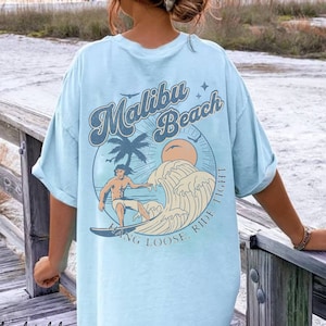 Puede incluir: Camiseta azul claro con un gráfico retro de un surfista montando una ola con el texto "Malibu Beach" y "Hang Loose, Ride Tight" en un círculo.