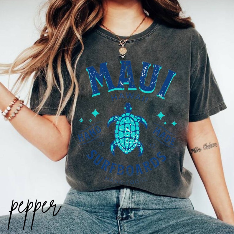 Pode incluir: Uma camiseta cinza escura com um design gr&aacute;fico azul e turquesa que apresenta o texto "MAUI HAWAII HAND MADE SURFBOARDS". O gr&aacute;fico tamb&eacute;m inclui uma tartaruga marinha estilizada.