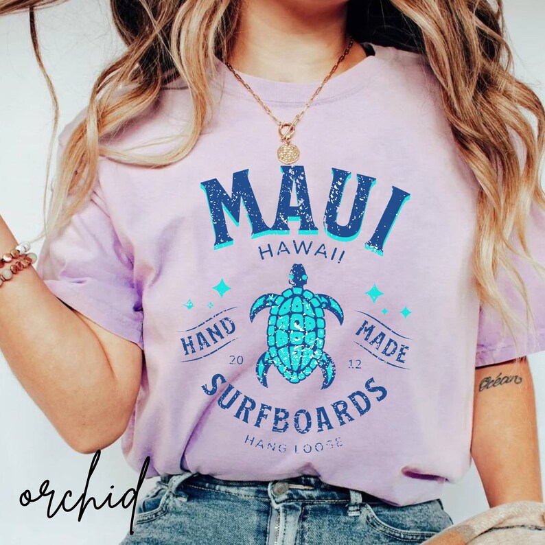 Pode incluir: Uma camiseta de cor lil&aacute;s claro com um gr&aacute;fico azul desbotado que diz "Maui Hawaii!" com uma tartaruga marinha azul e o texto "Hand Made Surfboards Hang Loose" abaixo.
