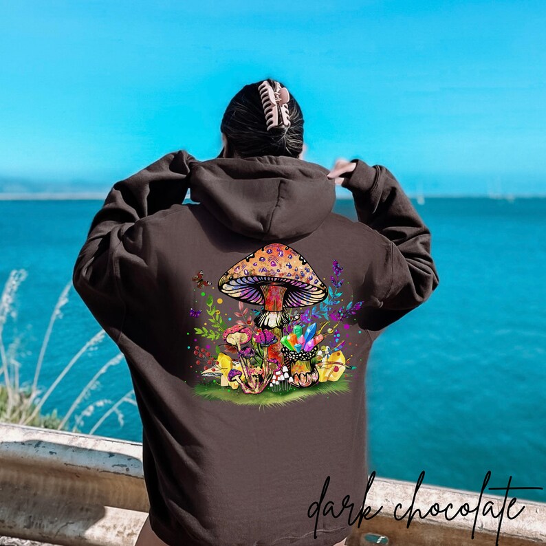 May include: A dark chocolate hoodie with a colorful mushroom and crystal graphic on the back. The mushroom has a purple and orange cap with white spots. The crystals are blue, purple, and green. The graphic is surrounded by green leaves and flowers.