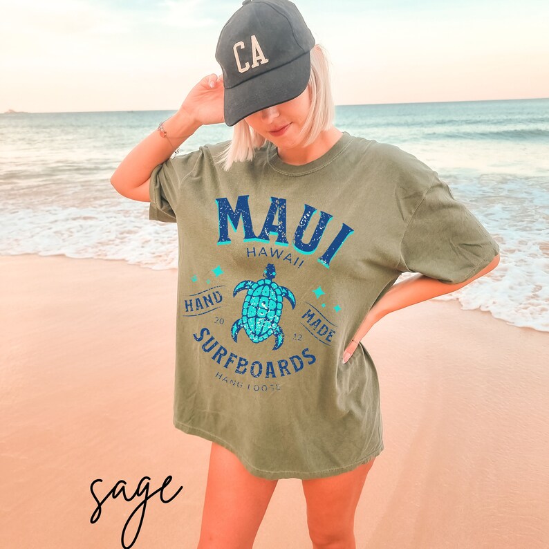 Pode incluir: Camiseta verde-s&aacute;lvia com um gr&aacute;fico de uma tartaruga marinha e o texto "Maui Hawaii Hand Made Surfboards Hand 100%".
