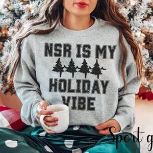 Könnte beinhalten: Ein hellgraues Sweatshirt mit dem Text "NSR IS MY HOLIDAY VIBE" in schwarzen Blockbuchstaben und vier Weihnachtsbäumen. Die Person hält einen weißen Becher. Die Worte "Sport grey" sind kursiv geschrieben.