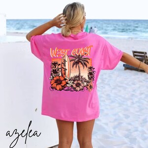 May include: Pink oversized t-shirt with a graphic print of a surfboard, palm tree, and flowers. The text "West Coast surf shop" is printed on the surfboard.