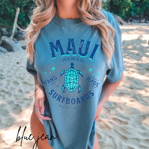 Pode incluir: Uma camiseta azul claro com um design gr&aacute;fico desbotado que apresenta o texto "Maui Hawaii" e uma tartaruga marinha turquesa. O gr&aacute;fico tamb&eacute;m inclui o texto "Hand Made Surfboards Hang 100".