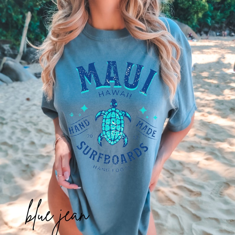 Pode incluir: Uma camiseta azul claro com um design gr&aacute;fico desbotado que apresenta o texto "Maui Hawaii" e uma tartaruga marinha turquesa. O gr&aacute;fico tamb&eacute;m inclui o texto "Hand Made Surfboards Hang 100".