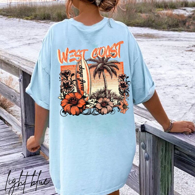 May include: Light blue t-shirt with a graphic print of a surfboard, palm trees, and flowers. The text "West Coast surf shop" is printed on the surfboard.