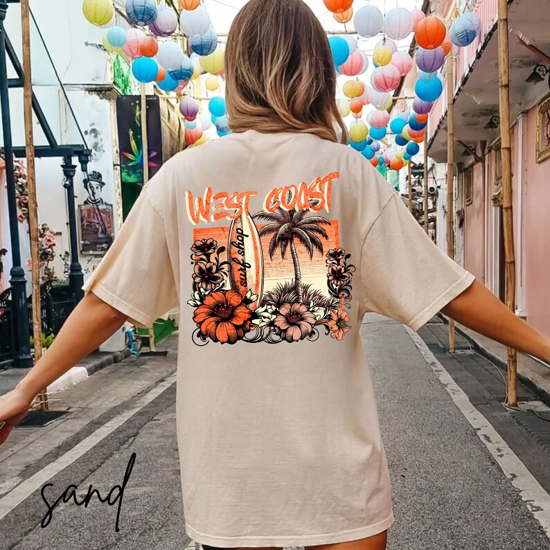 May include: A beige t-shirt with a graphic print of a palm tree, surfboard, and flowers. The text "West Coast surf shop" is printed on the shirt.