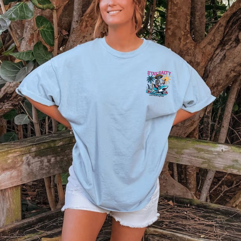 May include: A light blue t-shirt with a graphic of a skeleton riding a shark with the text "Stay Salty" and "Ocean Beach Salty Life" printed on the front.