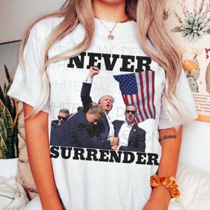 May include: White t-shirt with a graphic print of a group of people standing in front of an American flag. The text "NEVER SURRENDER" is printed in black letters on the shirt.
