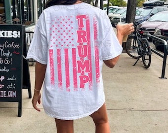 May include: White t-shirt with a pink American flag design and the word "TRUMP" printed vertically down the flag.