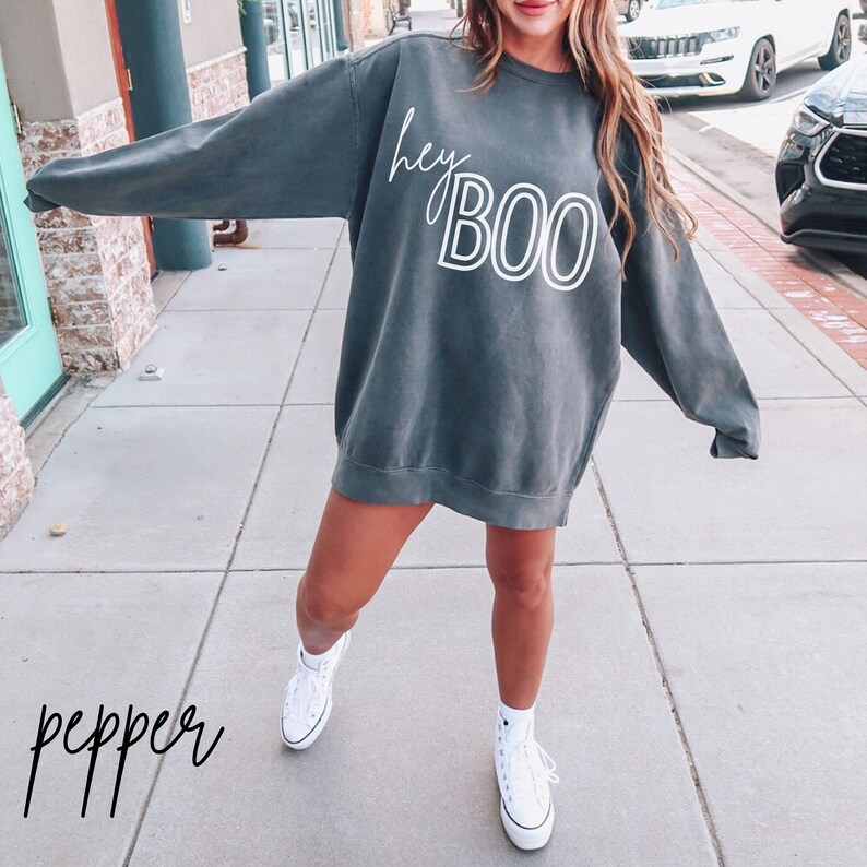 May include: A gray sweatshirt with the text "hey boo" in white lettering. The sweatshirt is worn by a person standing on a sidewalk.