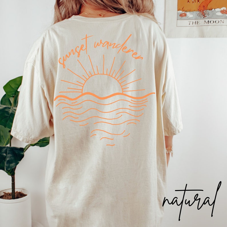 May include: A beige t-shirt with an orange graphic of a sunset over the ocean and the text "sunset wanderers". The text "natural" is written in black at the bottom of the shirt.