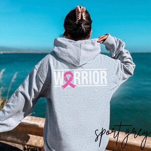 May include: A gray hooded sweatshirt with the word "WARRIOR" printed in white on the back. A pink ribbon with a white outline is centered above the word.