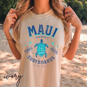Pode incluir: Uma camiseta bege claro com um design gr&aacute;fico azul e turquesa. O gr&aacute;fico apresenta o texto "MAUI HAWAII" com uma tartaruga e o texto "HAND MADE SURFBOARDS HANG LOOSE".