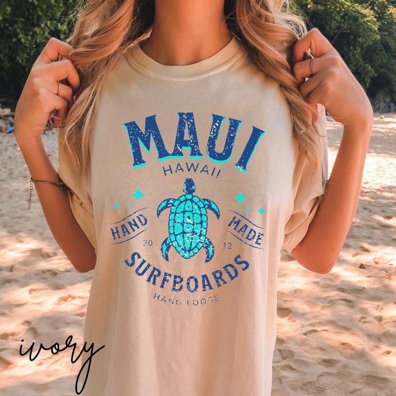 Pode incluir: Uma camiseta bege claro com um design gr&aacute;fico azul e turquesa. O gr&aacute;fico apresenta o texto "MAUI HAWAII" com uma tartaruga e o texto "HAND MADE SURFBOARDS HANG LOOSE".