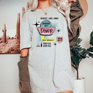 以下が含まれることがあります： レトロなダイナーの看板デザインが特徴のライトグレーのTシャツ。「Diner」の文字と「Breakfast, Lunch, Dinner」の文字がピンクとターコイズブルーで描かれています。「Emergency Dept.」、「Daily Specials」、「Turkey Club」、「Cherry Cola」、「Open 24/7」の文字も含まれています。