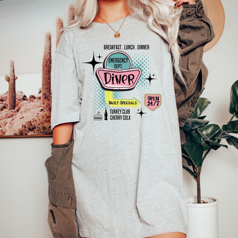 以下が含まれることがあります： レトロなダイナーの看板デザインが特徴のライトグレーのTシャツ。「Diner」の文字と「Breakfast, Lunch, Dinner」の文字がピンクとターコイズブルーで描かれています。「Emergency Dept.」、「Daily Specials」、「Turkey Club」、「Cherry Cola」、「Open 24/7」の文字も含まれています。
