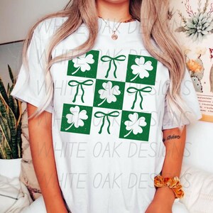 以下が含まれることがあります： 緑のクローバーとボウのデザインが施された白いTシャツ。デザインはクローバーとボウの市松模様です。