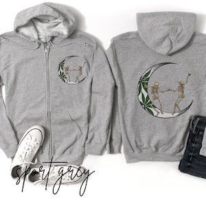 May include: Two skeletons dancing on a crescent moon with a marijuana leaf design. The skeletons are wearing a white shirt and black pants. The moon is white with a green marijuana leaf design. The image is on a gray hoodie.