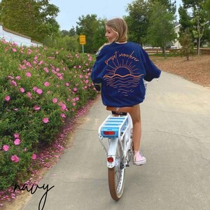 May include: A navy blue sweatshirt with the text "sunset wanderer" and a graphic of a sunset over the ocean. The sweatshirt is being worn by a person riding a bicycle.