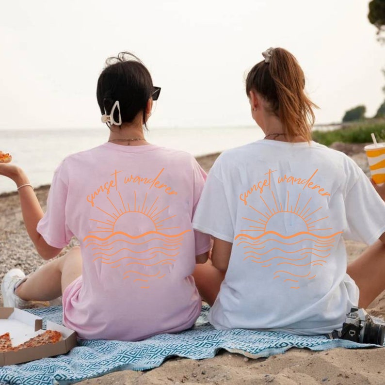 May include: Two people wearing matching t-shirts with the text "sunset wanderers" and a graphic of a sunset over the ocean. The shirts are pink and white.