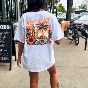 May include: White t-shirt with a graphic print of a surfboard, palm trees, and flowers. The text "WEST COAST" and "surf shop" are printed on the shirt.
