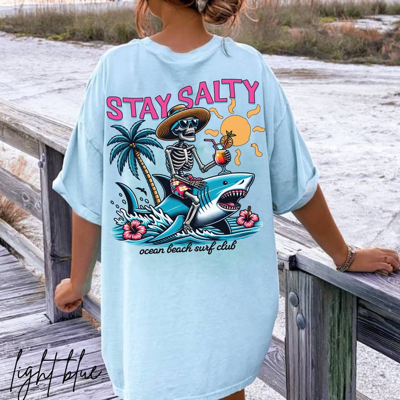 May include: Light blue t-shirt with a graphic of a skeleton riding a shark and holding a drink. The text "STAY SALTY" is printed in pink on the shirt. The text "ocean beach surf club" is printed below the graphic.