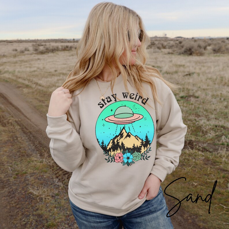 May include: A beige sweatshirt with a graphic of a flying saucer over a mountain range with a forest and flowers. The text "stay weird" is above the graphic.