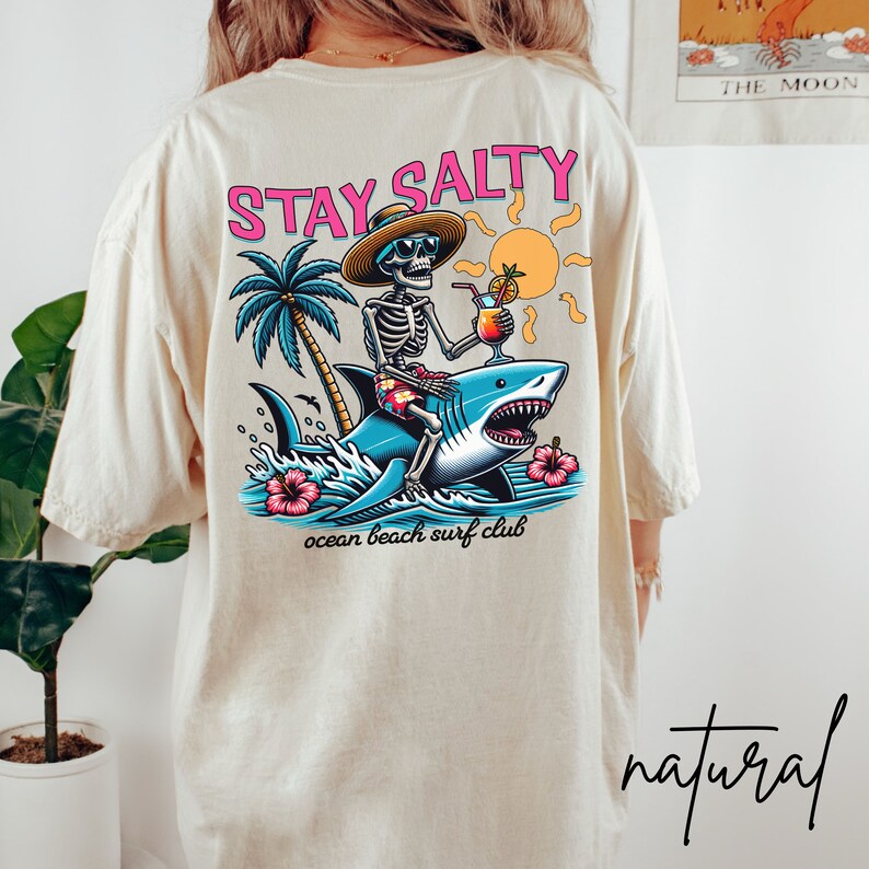 May include: A white t-shirt with a graphic print of a skeleton riding a shark. The graphic includes a palm tree, a sun, and the text "Stay Salty" and "ocean beach surf club".