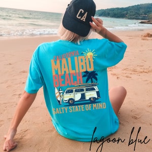 May include: A turquoise blue t-shirt with a graphic print of a vintage van with a surfboard on top. The graphic also includes a palm tree, a sun, and the text "California Malibu Beach Salty State of Mind".