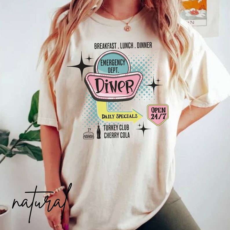 以下が含まれることがあります： レトロなダイナーのデザインが施されたクリーム色のTシャツ。グラフィックには、「Diner」の文字が入ったピンクとティールブルーのダイナーの看板があり、「Breakfast, Lunch, Dinner」、「Daily Specials」、「Open 24/7」の文字が囲んでいます。