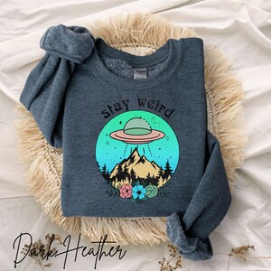 May include: Dark heather grey sweatshirt with a graphic of a UFO flying over a mountain range with a forest in the foreground. The graphic is surrounded by a circle with the text "Stay Weird" above the graphic.