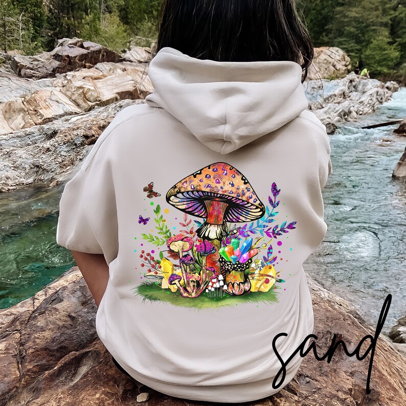 May include: A beige hoodie with a colorful mushroom graphic on the back. The mushroom is surrounded by flowers and butterflies. The text "sand" is printed on the bottom right corner of the hoodie.