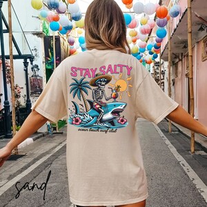 May include: A beige t-shirt with a graphic of a skeleton riding a shark. The graphic includes palm trees, a sun, and the text "STAY SALTY" in pink. The text "ocean beach surf club" is below the graphic.
