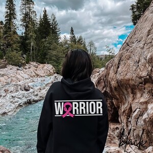 May include: Black hoodie with a white "Warrior" text and a pink ribbon graphic on the back. The hoodie is worn by a person standing in front of a rocky landscape.