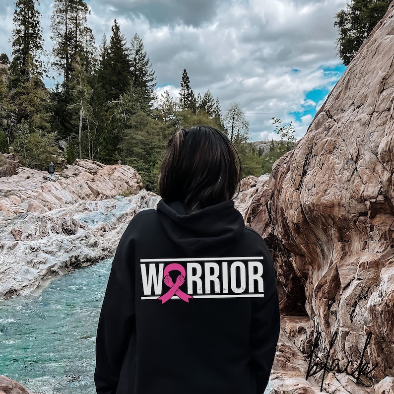 May include: Black hoodie with a white "Warrior" text and a pink ribbon graphic on the back. The hoodie is worn by a person standing in front of a rocky landscape.